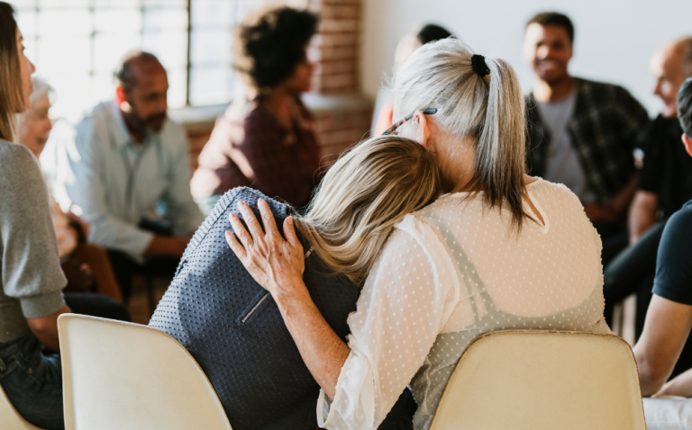 Compass Associates - A group of people within a room sat in a circle as if portraying a support group blurred in the background. The image focusses on two people sat next to each other hugging in a supportive manner.