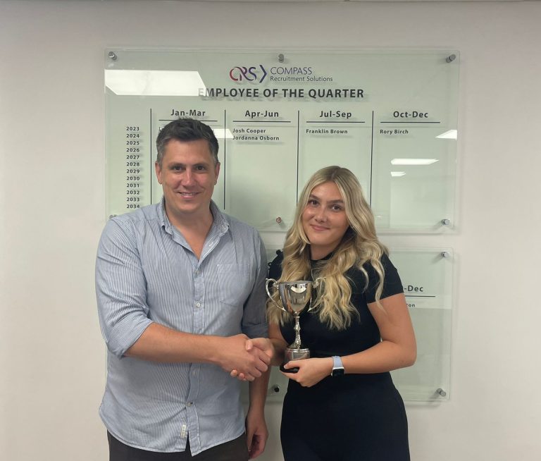 Compass Associates - Sam Leighton-Smith (A male) and Jordanna Osborn (A female) are standing in front of a wall-mounted board that reads "Employee of the Quarter." The board lists names of award recipients under the months of Jan-Mar, Apr-Jun, Jul-Sep, and Oct-Dec for the years 2023 to 2034. Sam is on the left, wearing a light blue button-up shirt and dark pants, and is shaking hands with the woman. Jordanna, on the right, is holding a trophy and is dressed in a black outfit with an Apple Watch on her wrist. Both are smiling at the camera.